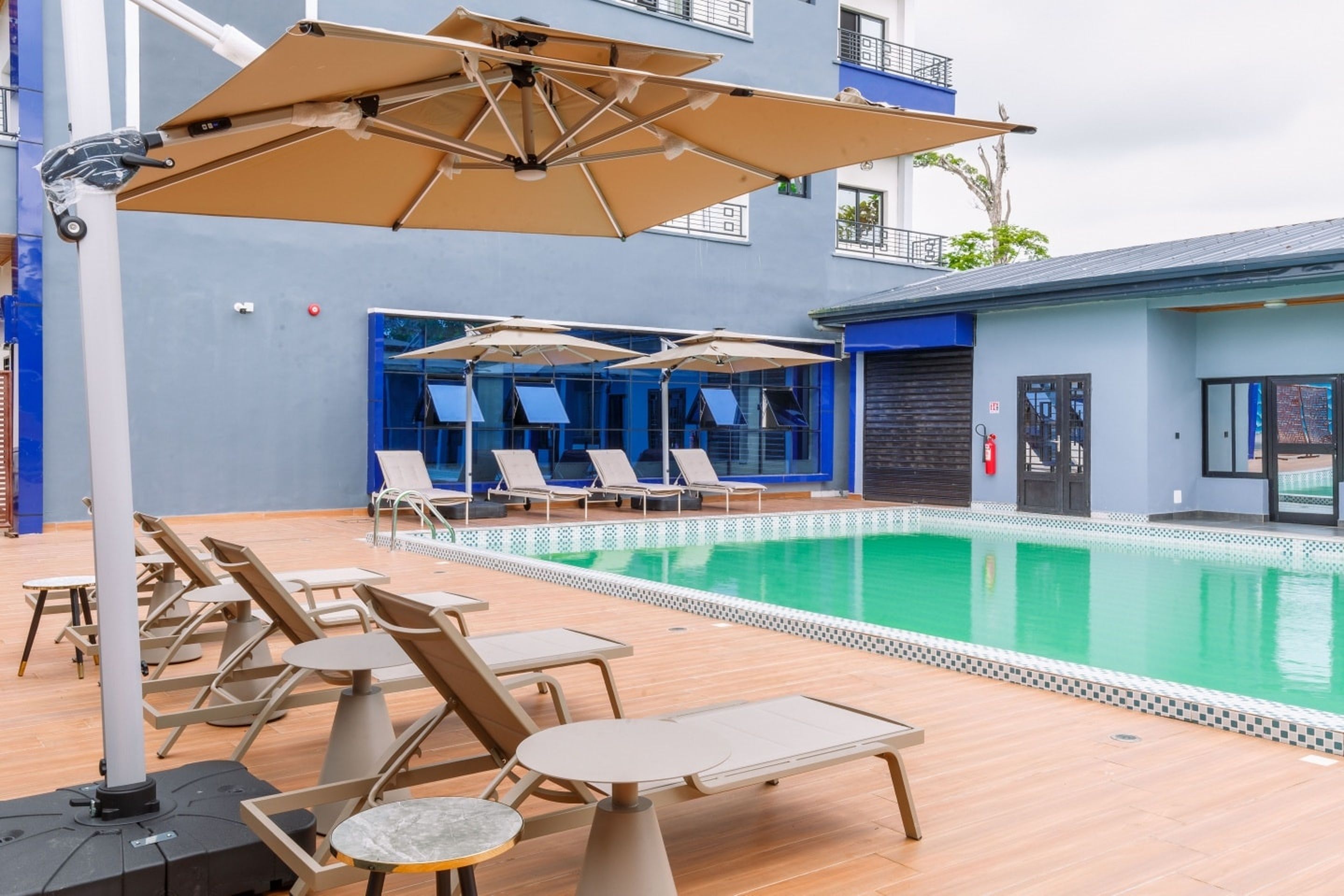 Zona de piscina con sillones de descanso y sombrillas, una piscina y un edificio con paredes exteriores azules y blancas.