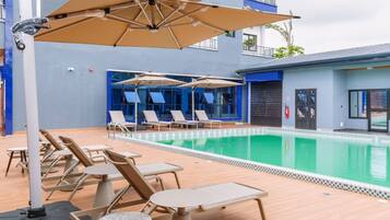 Zona de piscina con sillones de descanso y sombrillas, una piscina y un edificio con paredes exteriores azules y blancas.