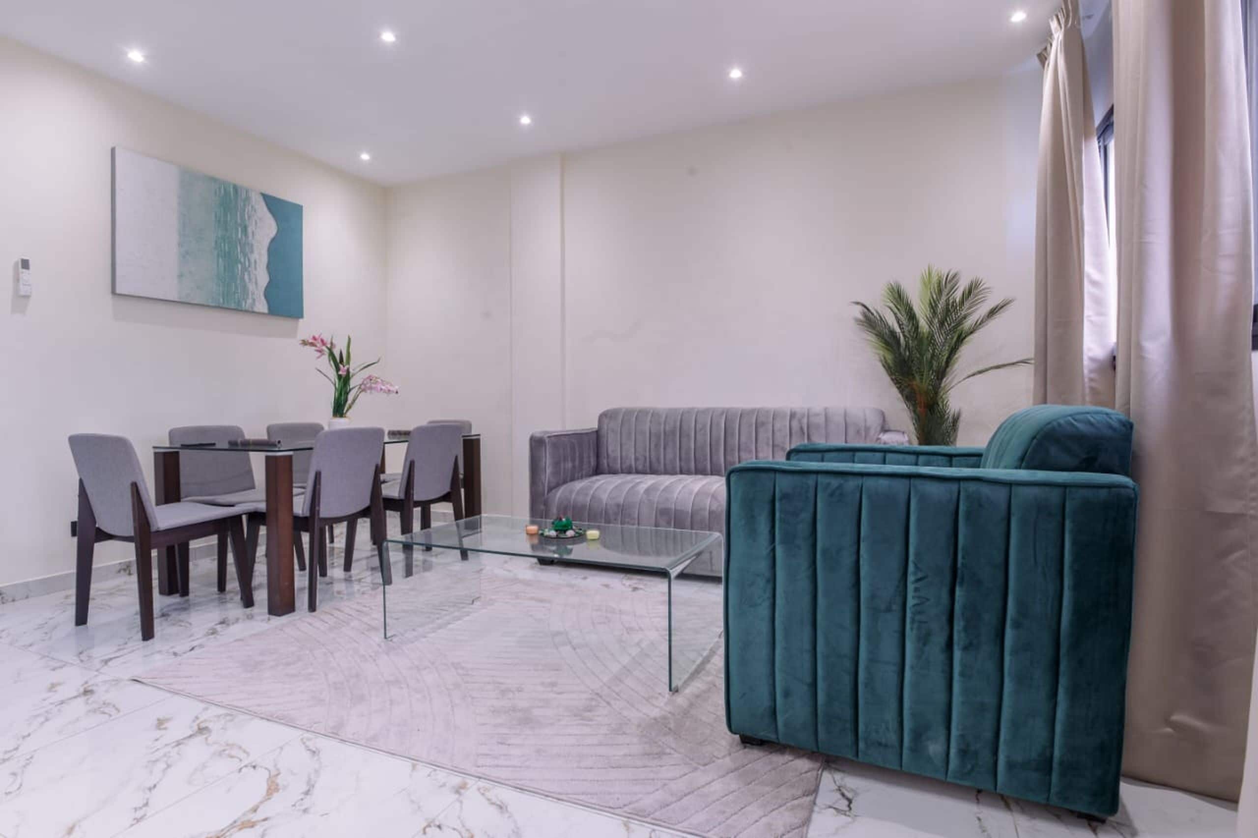Uma sala de estar moderna com mesa de centro de vidro, sofá cinza, poltrona verde-água e uma área de jantar com cadeiras e mesa.