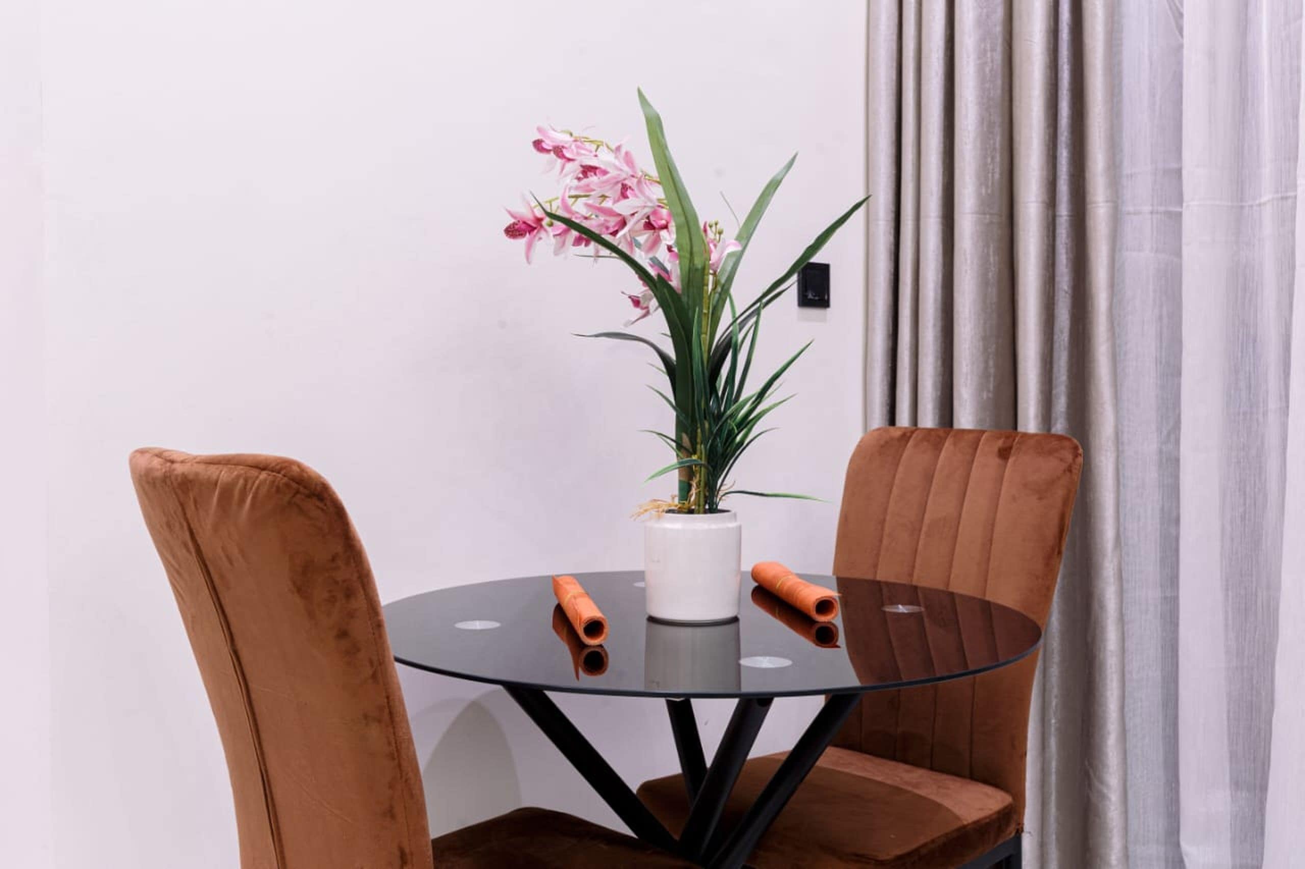 A round glass table with two brown upholstered chairs, a potted plant with pink flowers, and a white vase.