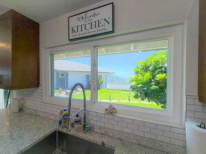 A kitchen with a window view of a house and greenery, a welcome sign, and a stainless steel sink. - Sweeping Ocean & Koko Head Views - Ridgetop Home (Honolulu)