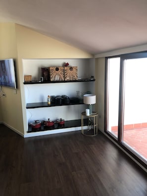 Ein modernes Wohnzimmer mit einer schwarzen Arbeitsplatte, Regalen mit Küchenutensilien, einem Fernseher und einem großen Fenster.