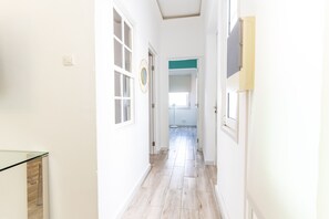 Ein schmaler Flur mit weißen Wänden und Holzdielen, der zu einem Zimmer mit Fenster und Tür führt.