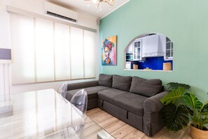 A modern living room with a sectional sofa, a dining table with chairs, a large window with blinds, a kitchen area with a microwave, and a large plant. - 3-Bedroom Smile Family Home (Los Llanos de Aridane)