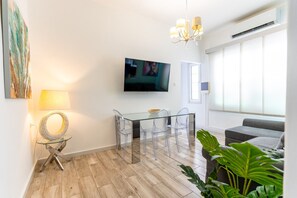 Ein modernes Wohnzimmer mit einem Glastisch, transparenten Stühlen, einem wandmontierten Fernseher, einer Kandelaberleuchte und einem grauen Sofa.