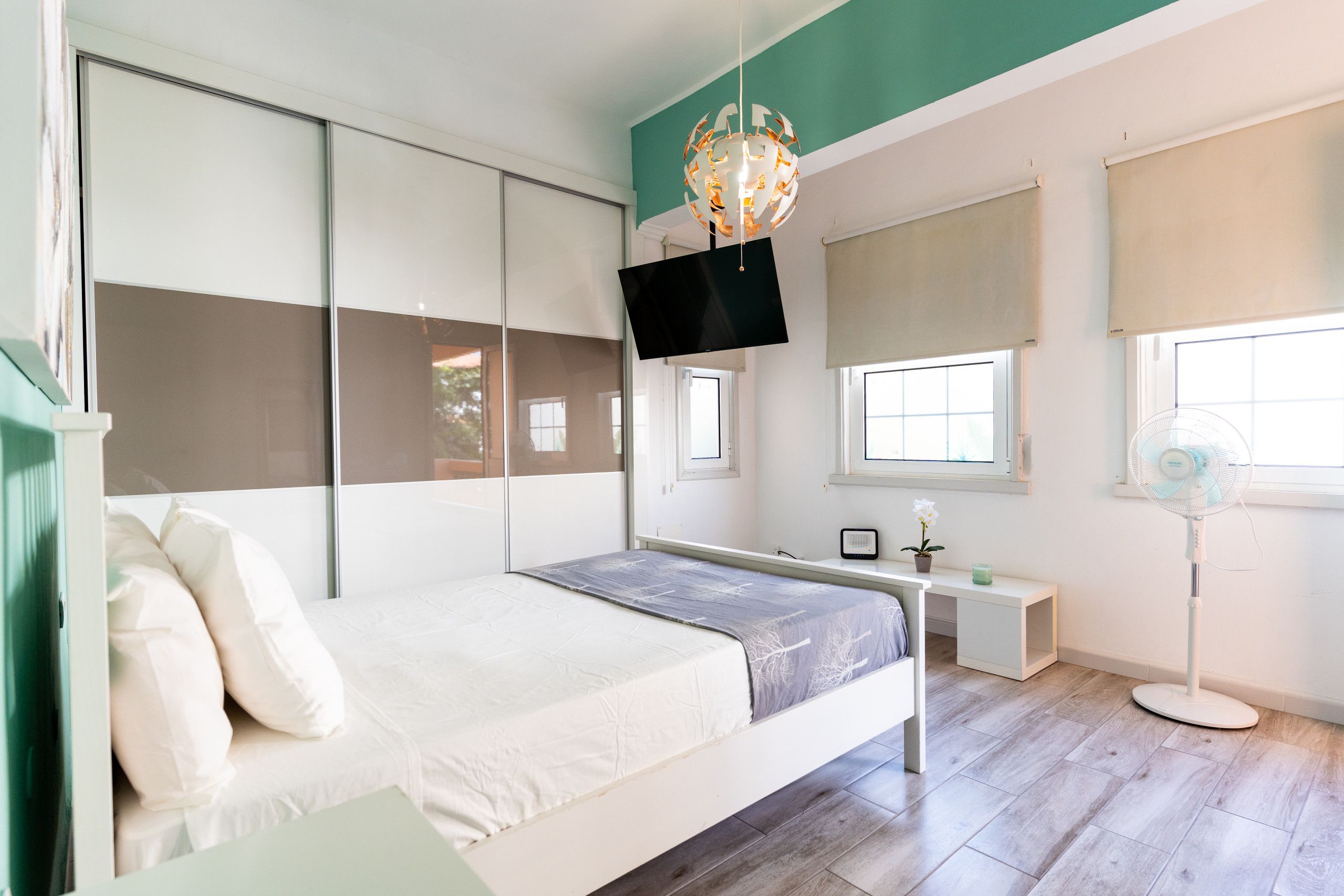 A modern bedroom with a bed, a desk, a TV, and a standing fan.