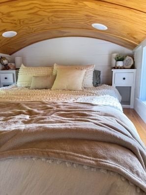 Un dormitorio con techo inclinado, una cama con colcha color beige, dos mesitas de noche con lámparas y una ventana.