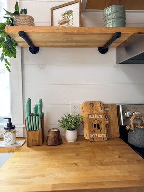 Una encimera de cocina con un estante de madera, una planta, un bloque de cuchillos, una vela, un frasco de jabón para manos y un letrero de madera.