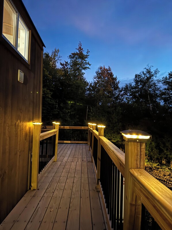 Una terraza de madera con barandas e iluminación, que conduce a un edificio con ventanas.