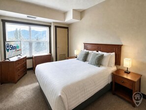 Ein Hotelzimmer mit einem großen Bett, zwei Nachttischen inklusive Lampen, einem Kleiderschrank, einem Fernseher und einem Fenster mit Jalousien.