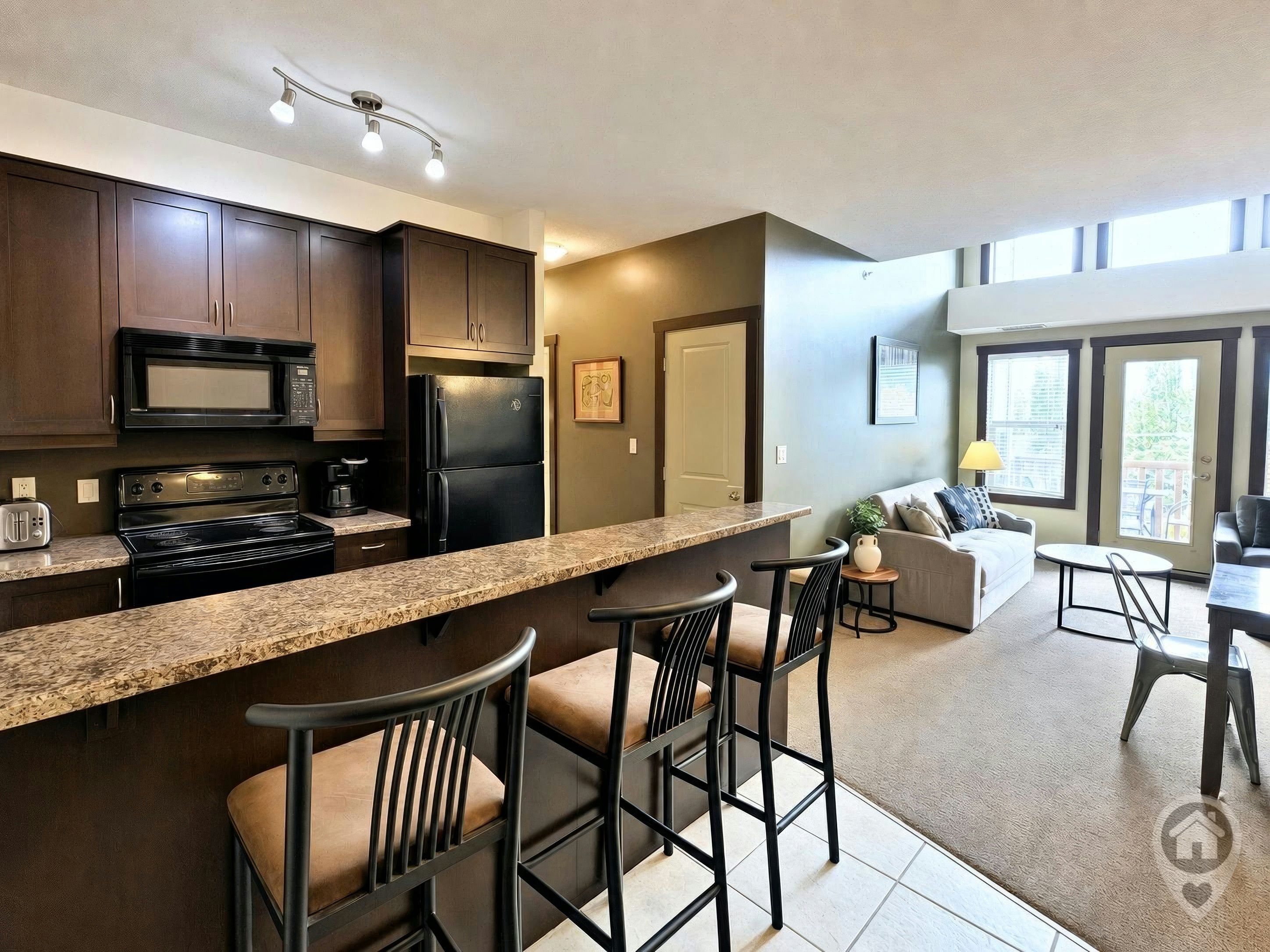 A modern kitchen with dark cabinets, a black refrigerator, and a microwave. There is a bar area with stools and a seating area with a sofa and a table.