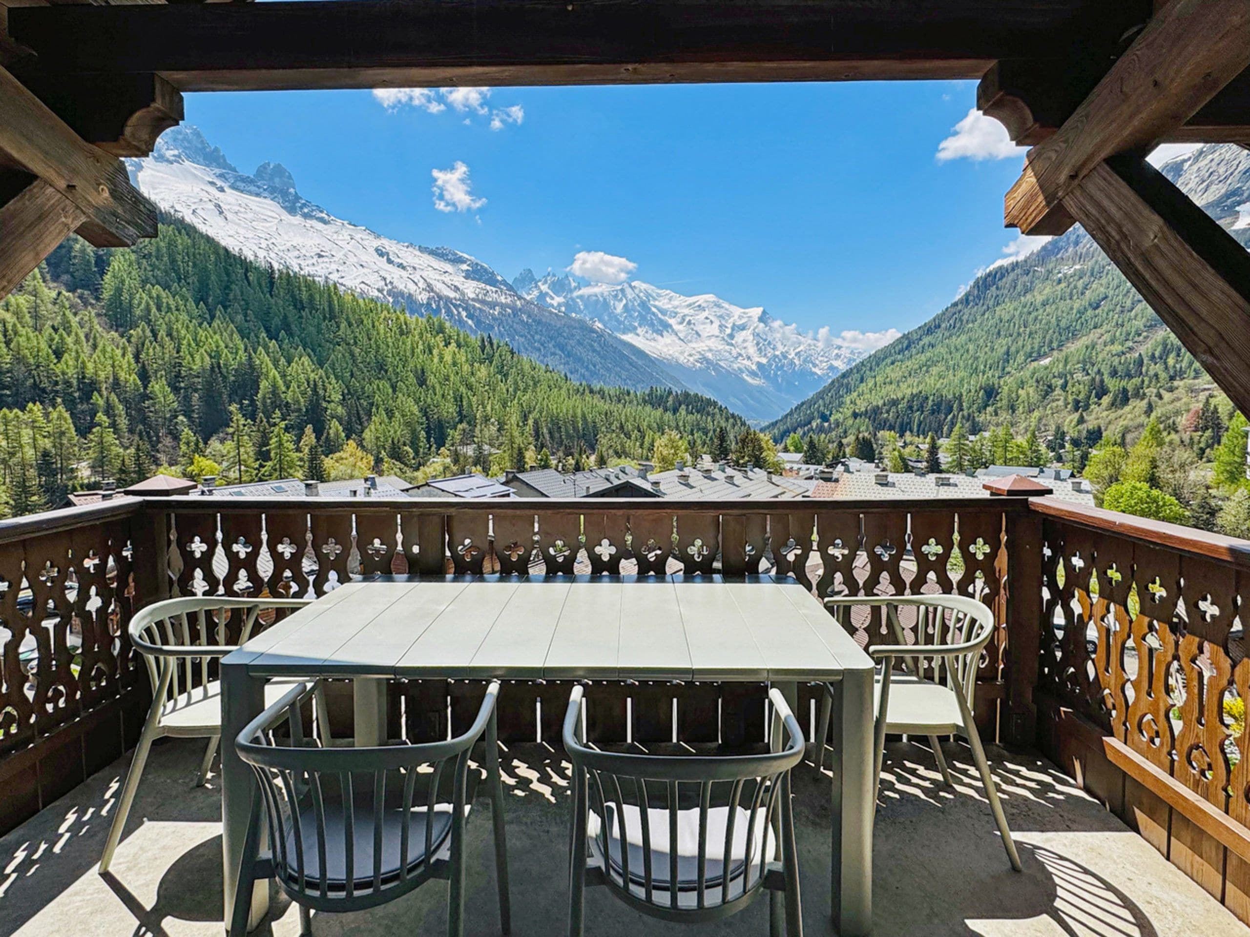 Eine Holzterrasse mit Tisch und Stühlen, mit Blick auf eine gebirgige Landschaft mit schneebedeckten Gipfeln und dichten Wäldern.
