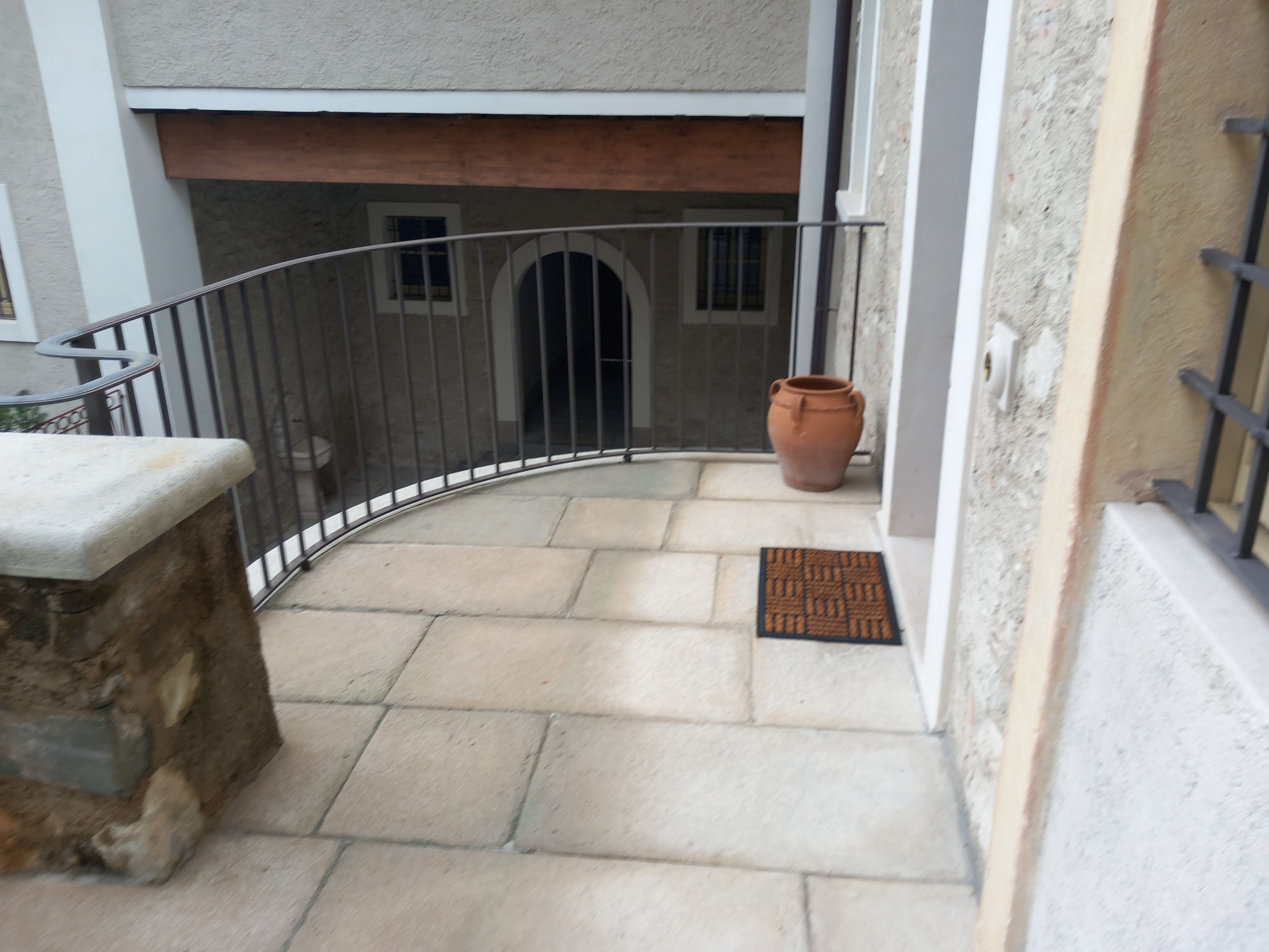 Un balcon incurvé avec une bordure en pierre, une rambarde en métal, un pot de fleurs et une porte avec un paillasson.