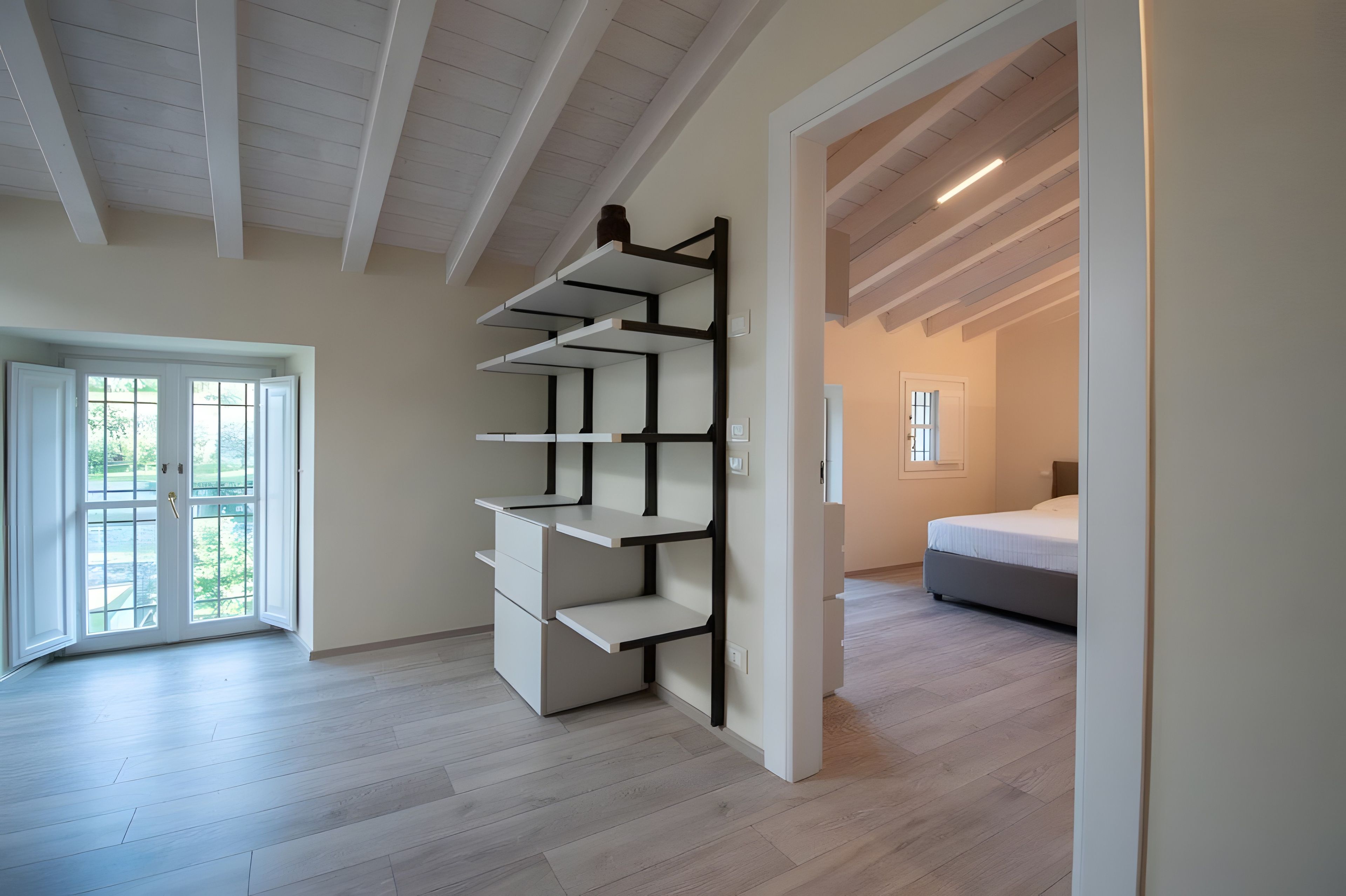 Ein Zimmer mit einer eingebauten Regallösung, einem Bett und Blick durch eine Tür auf einen Balkon.
