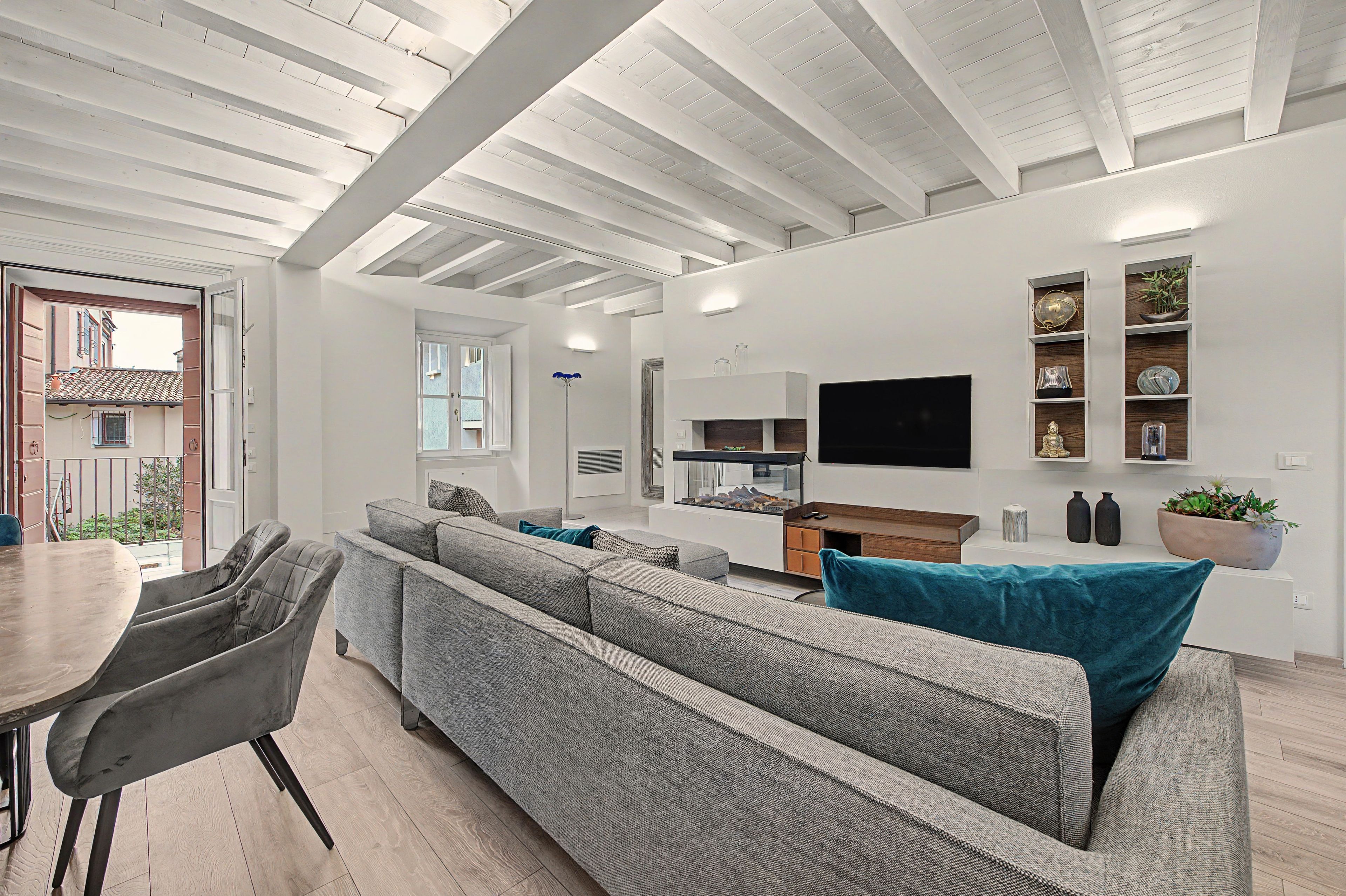 A modern living room with a large grey sofa, a flat-screen TV mounted on the wall, and a dining area with a table and chairs.