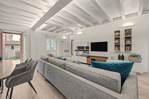 A modern living room with a large grey sofa, a flat-screen TV mounted on the wall, and a dining area with a table and chairs.