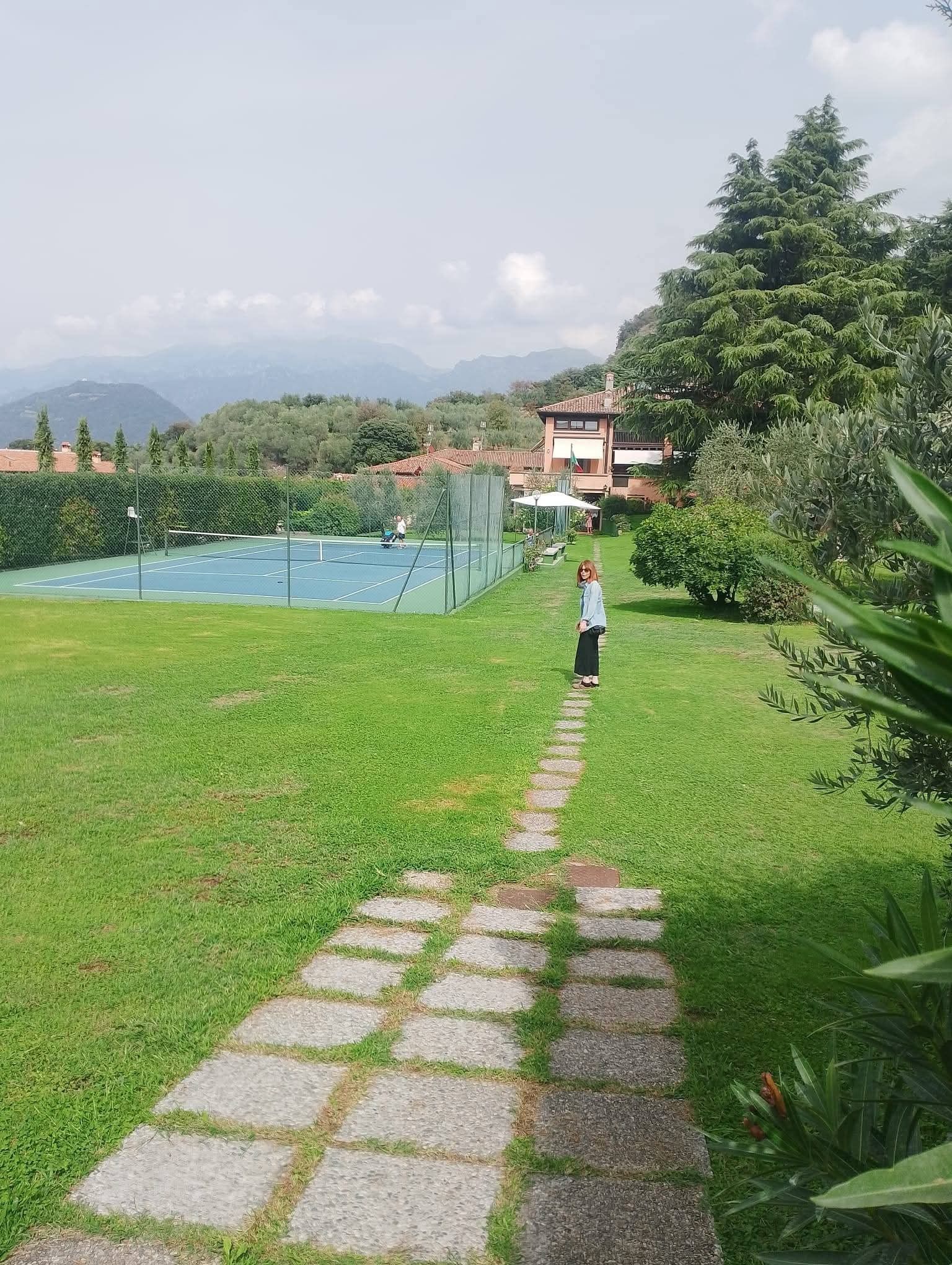A tennis court with a net, a stone pathway leading to a building, and a mountainous backdrop.