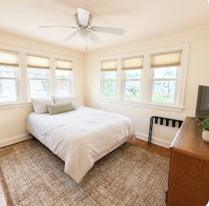 A bedroom with a bed, a ceiling fan, a radiator, and a TV on a wooden cabinet.