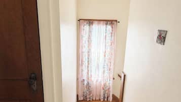 Um corredor estreito com soalho de madeira, uma porta de madeira à esquerda e uma janela com cortinas florais no final.