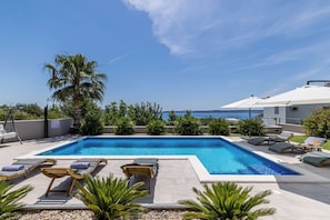A rectangular swimming pool with lounge chairs and umbrellas, surrounded by palm trees and a view of the sea. - Villa Belvedere, Sea View with Pool (Kaštel Kambelovac)