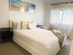 A neatly arranged bedroom with a bed, pillows, a bedside table, a lamp, a window with curtains, and a painting on the wall. - 42 Easy 2BR Business Stay Parking, Kitchen & Wi-Fi (Coogee)