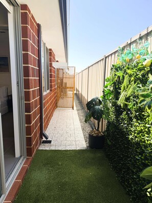 A narrow outdoor corridor with artificial grass, a brick wall, a metal gate, and dense foliage. - 42 Easy 2BR Business Stay Parking, Kitchen & Wi-Fi (Coogee)
