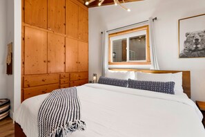 A bedroom with a wooden wardrobe, a bed with white bedding, a patterned throw, a window with curtains, and a bear artwork on the wall. - Big Bear Lake 2BR Cabin | Hot Tub, Arcade Game Room, Fire Pit & Covered Deck (Big Bear Lake)
