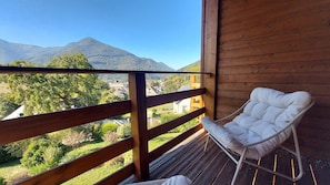 Een houten balkon met een witte stoel met kussens, met uitzicht op een bergachtig landschap met gebouwen in de verte.