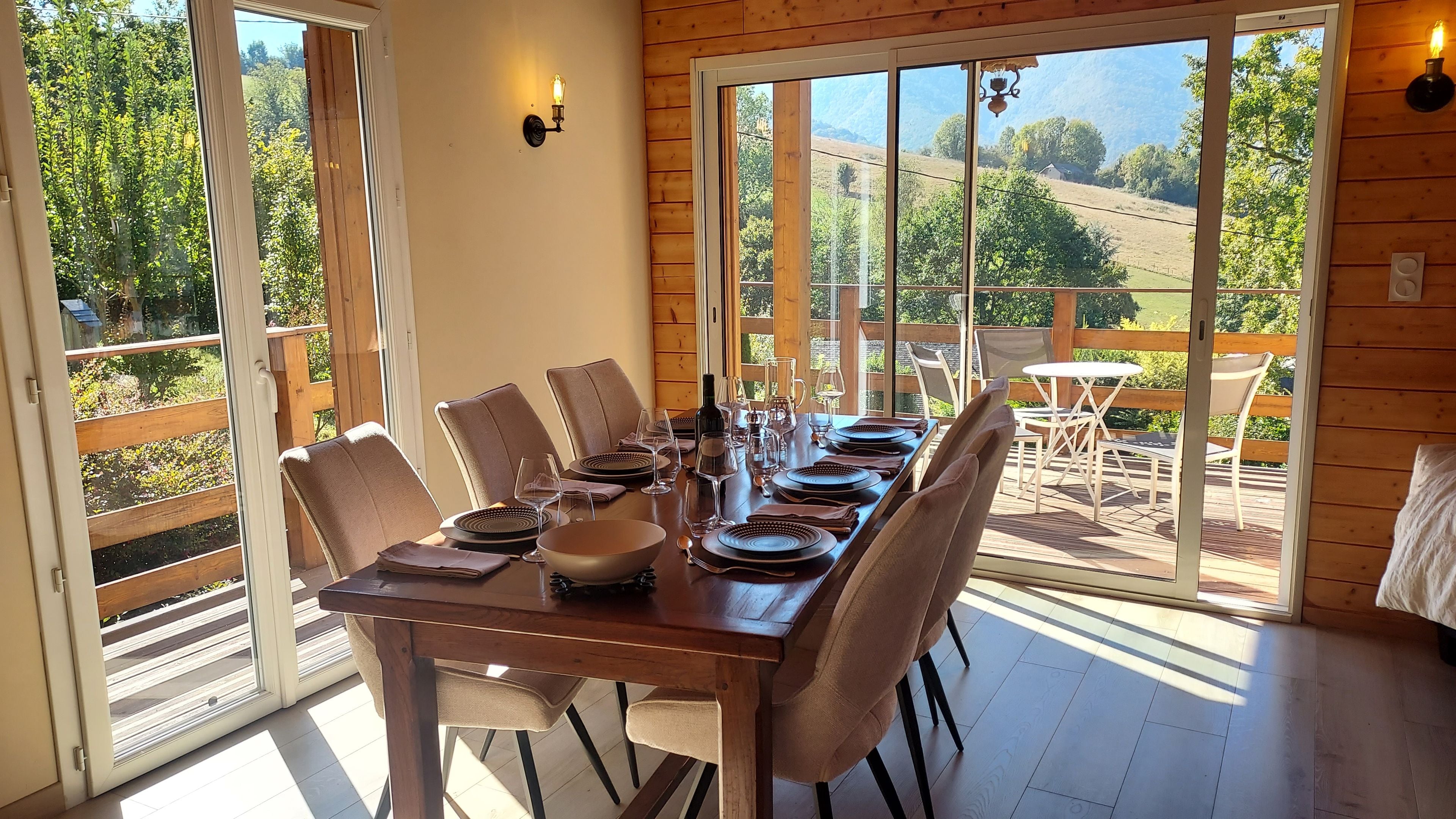 Un coin repas avec une table en bois dressée pour six personnes, entourée de chaises, et offrant une vue sur un jardin et des collines grâce à de grandes fenêtres.