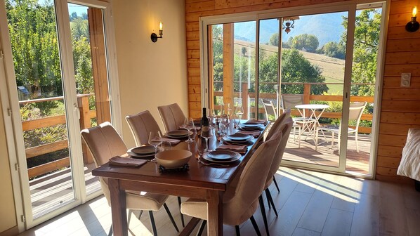 A dining area with a wooden table set for six, surrounded by chairs, with a view of a garden and hills through large windows.