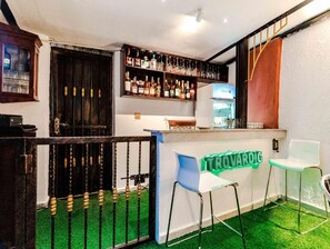 Un área de bar con un letrero de neón, estantes llenos de botellas, un refrigerador y dos taburetes.