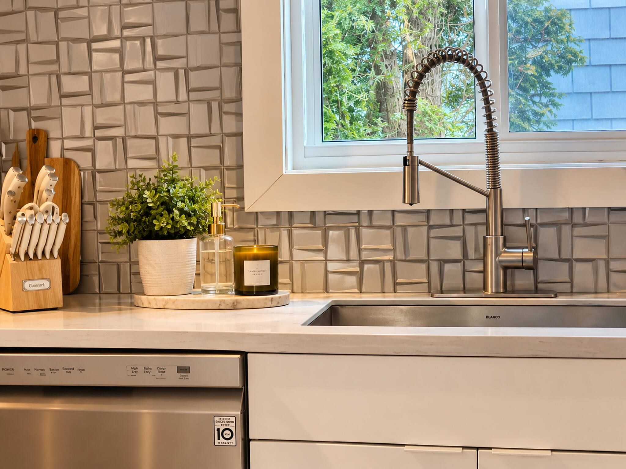 A modern kitchen with a built-in dishwasher, a stainless steel sink, and a window with a view of trees.