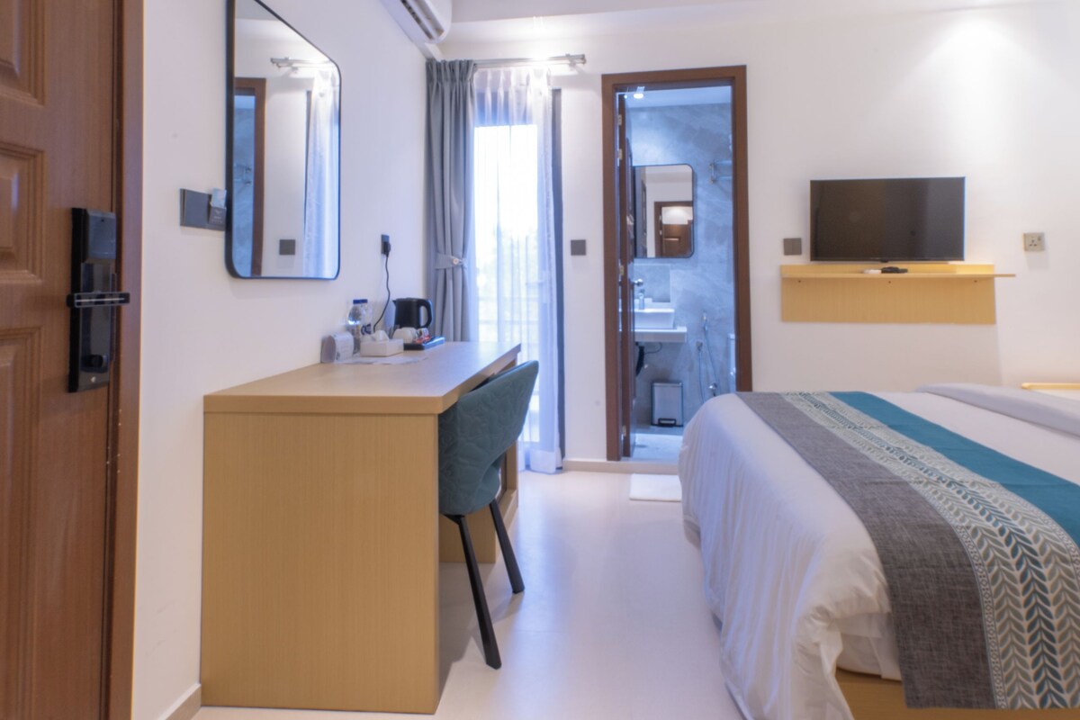 Une chambre d’hôtel équipée d’un lit, d’un bureau, d’une chaise, d’une télévision, d’un ventilateur de plafond et d’une salle de bain visible à travers une porte ouverte.