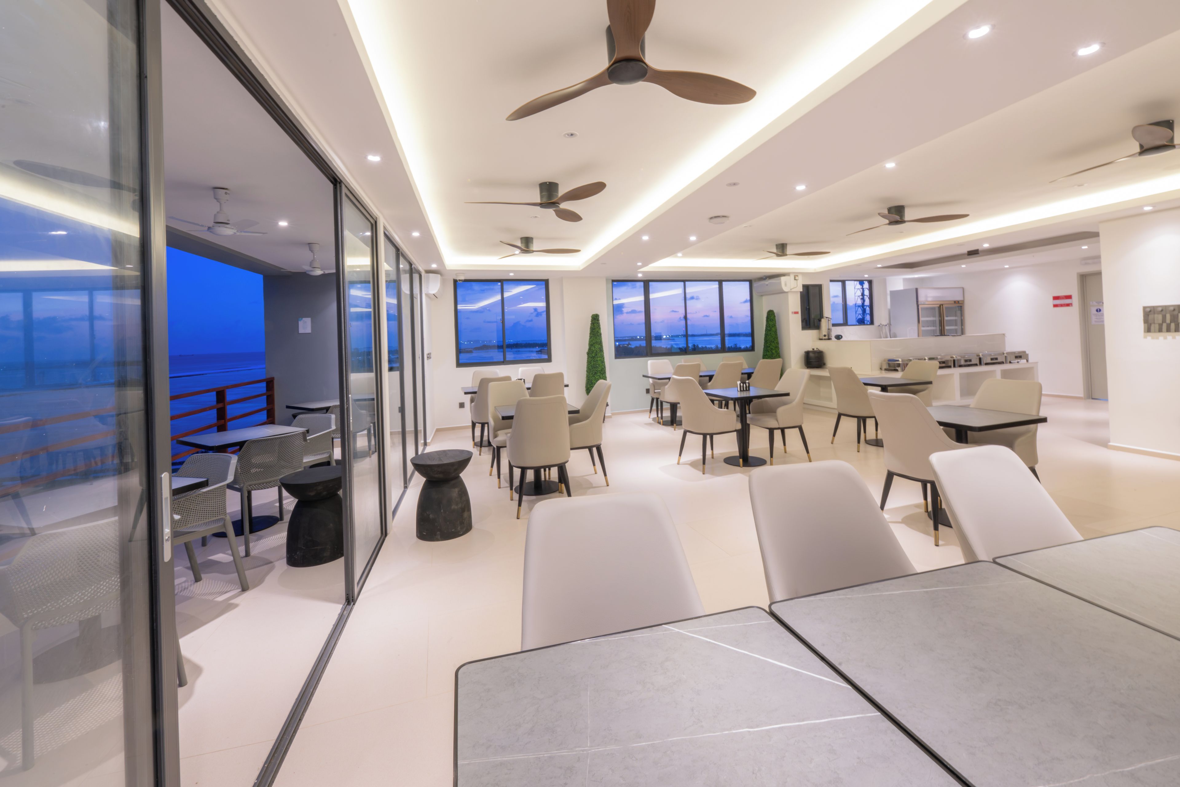 A modern dining area with tables and chairs, large windows with a view, and ceiling fans.
