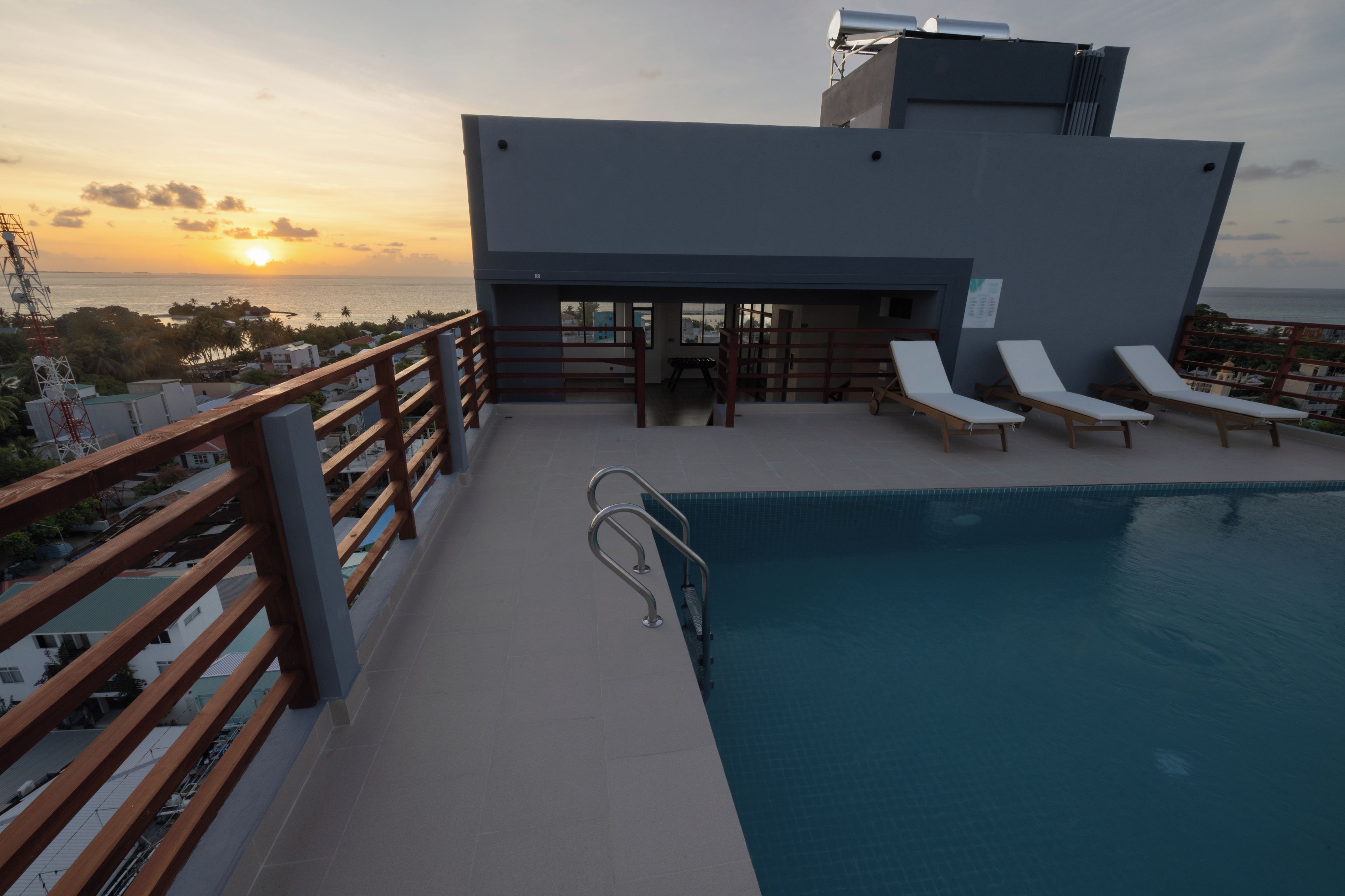 A rooftop pool area with a view of the sunset over the ocean, equipped with sun loungers and a metal pool ladder.