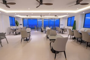 Une salle de conférence moderne, dotée de plusieurs tables et chaises, de grandes fenêtres offrant une vue sur la mer, de ventilateurs de plafond et d’un éclairage encastré.