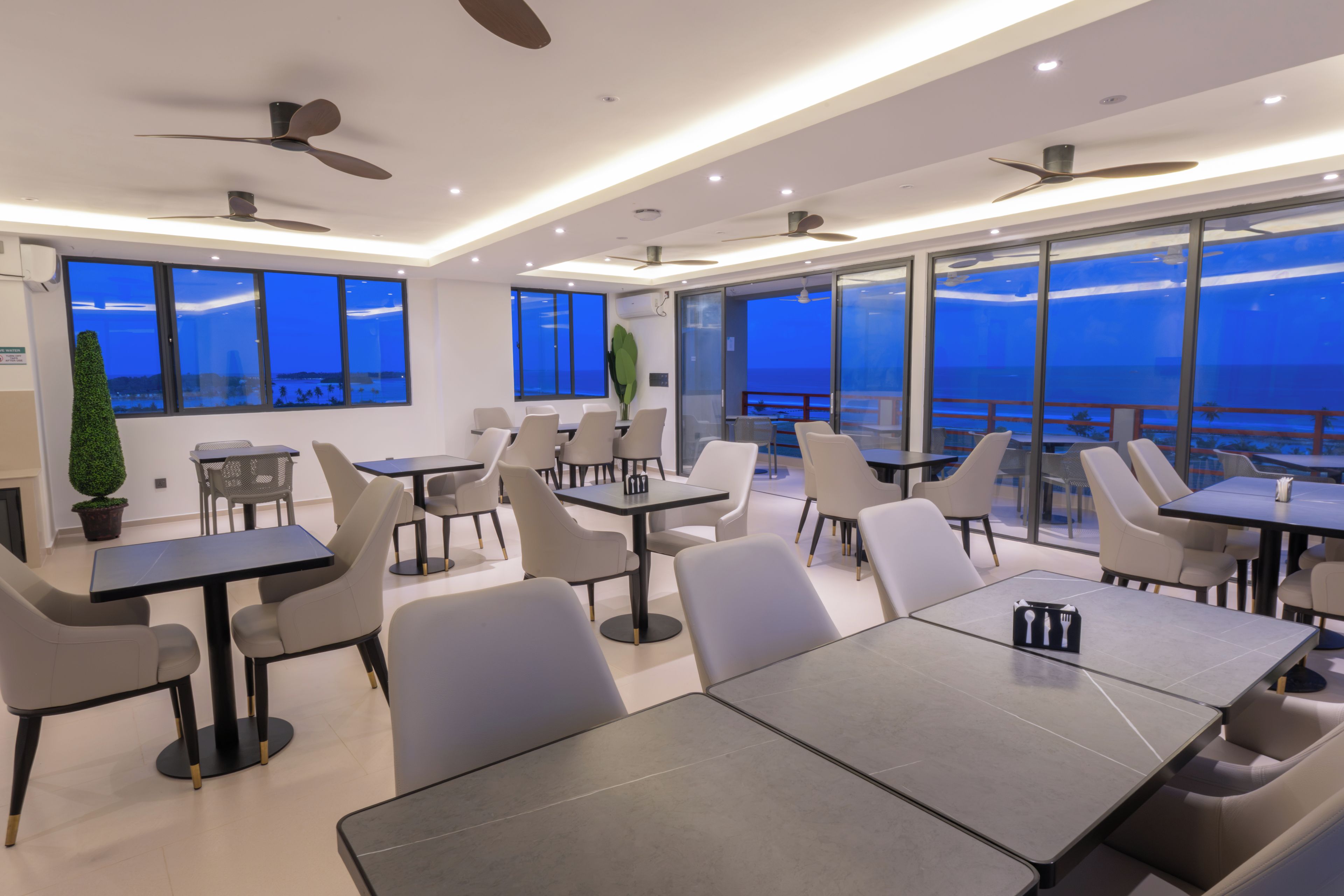 A modern dining area with tables and chairs, large windows with a view, and ceiling fans.