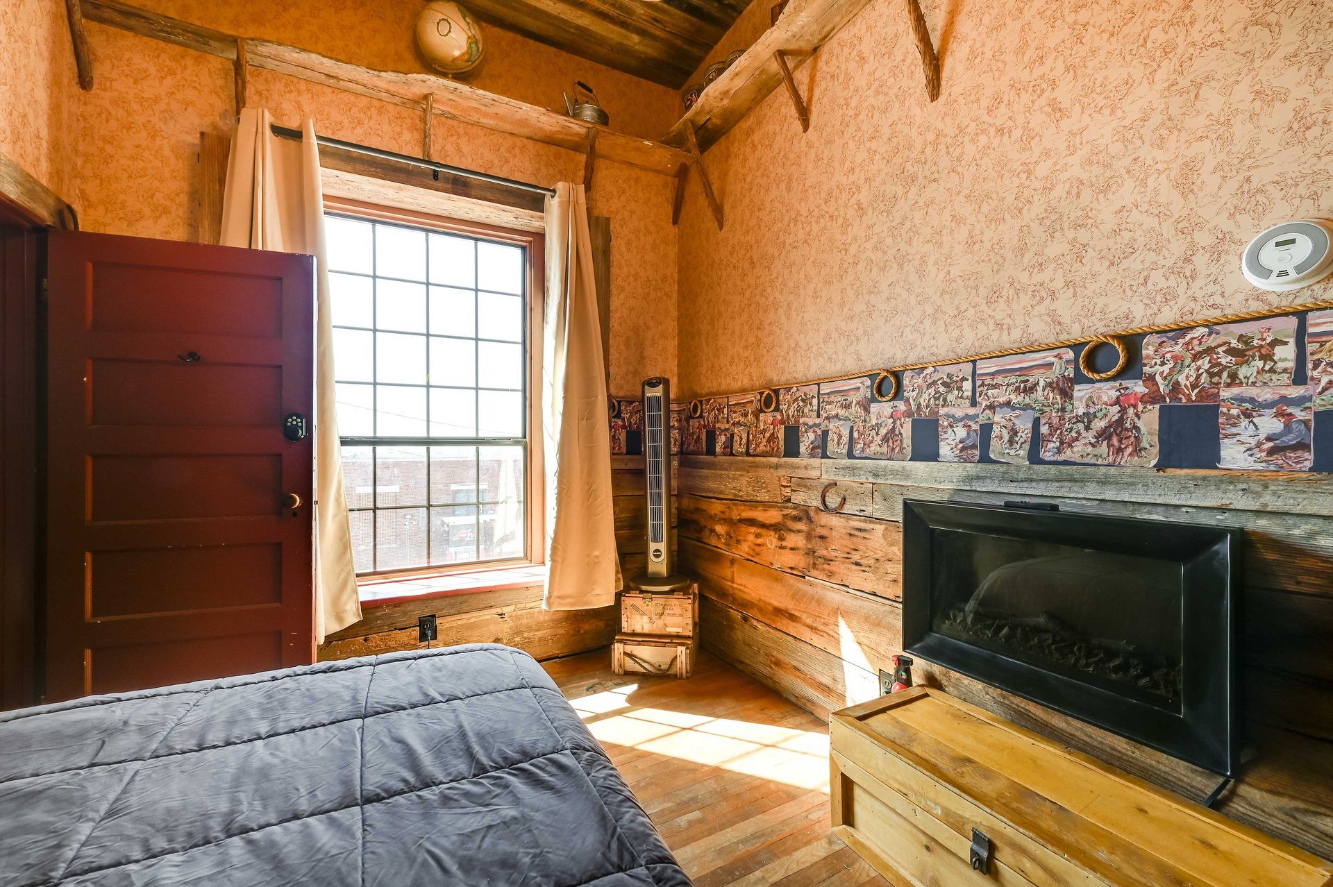 A room with a wooden floor, a bed, a fireplace, a window, and a wall with a decorative tile border.