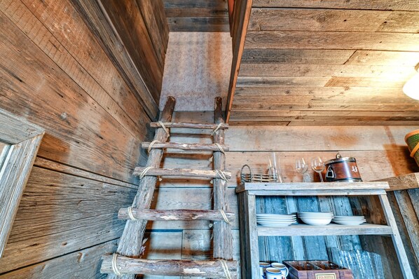 A rustic wooden interior with a ladder leading to a loft area, shelves with dishes and glasses, and a vintage-style metal container.