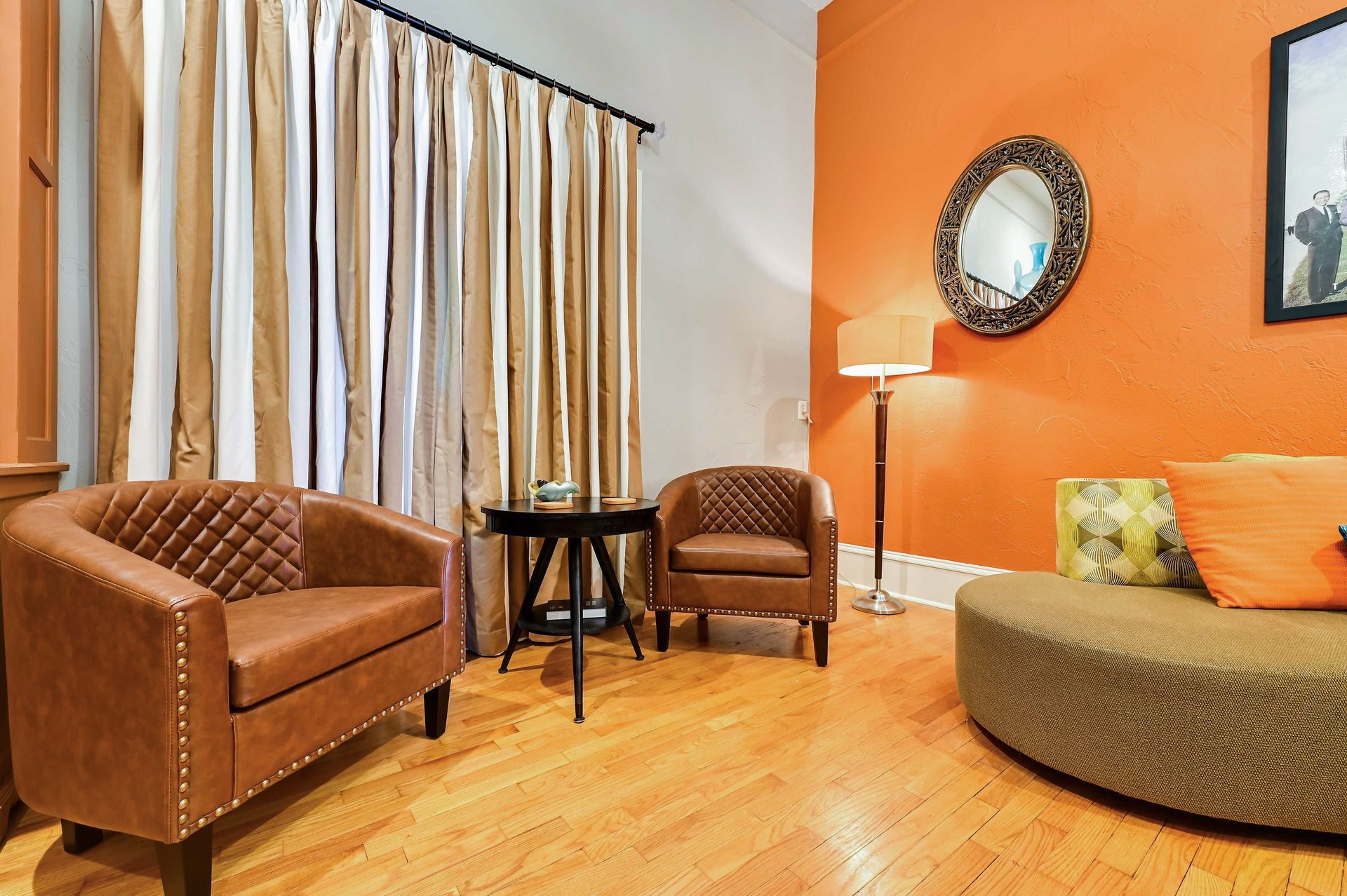 A room with a brown leather armchair, a smaller armchair, a round table, a floor lamp, and a sofa with a cushion.