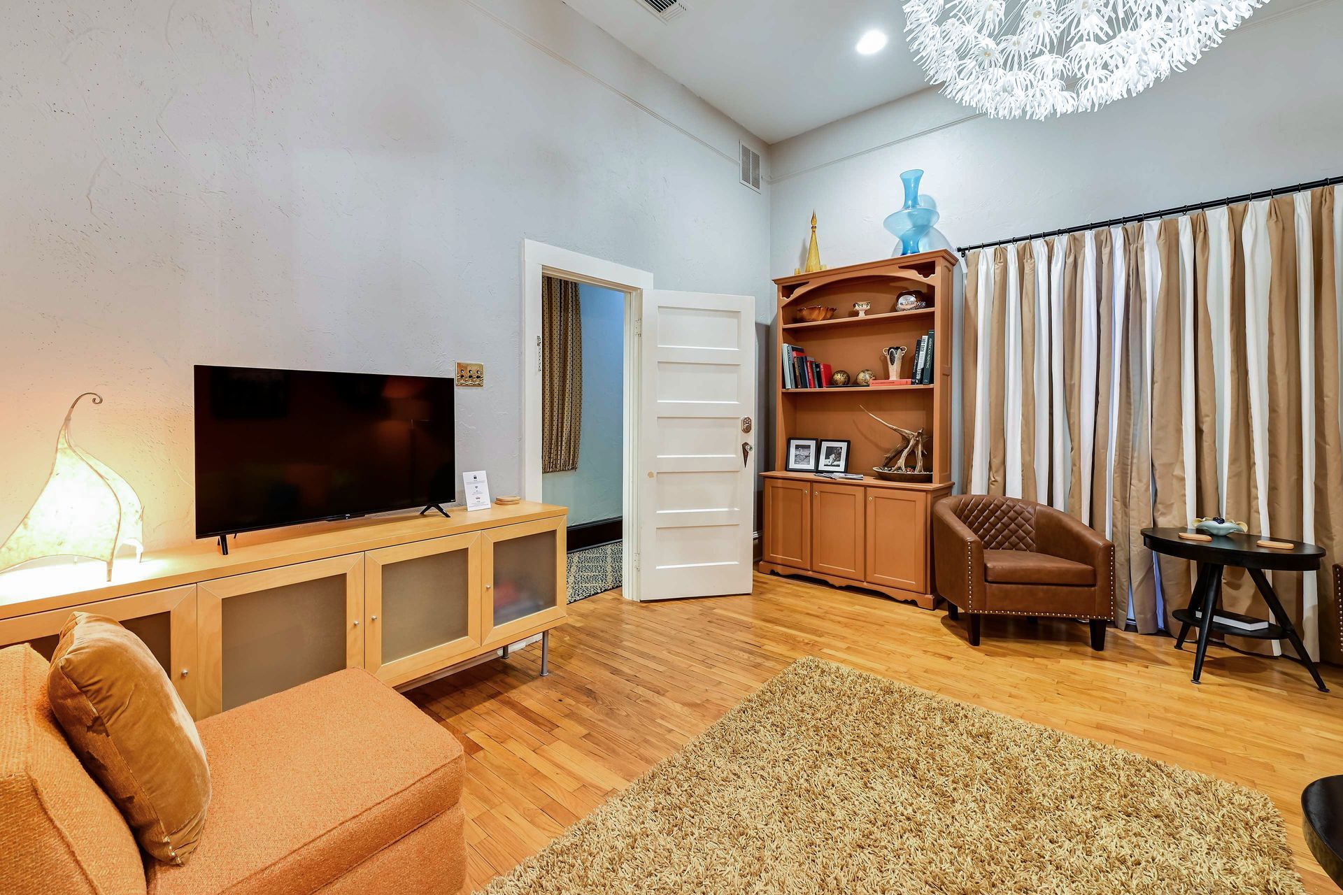 A living room with a flat-screen TV on a wooden stand, a brown leather armchair, a small round table, and a bookshelf with decorative items.