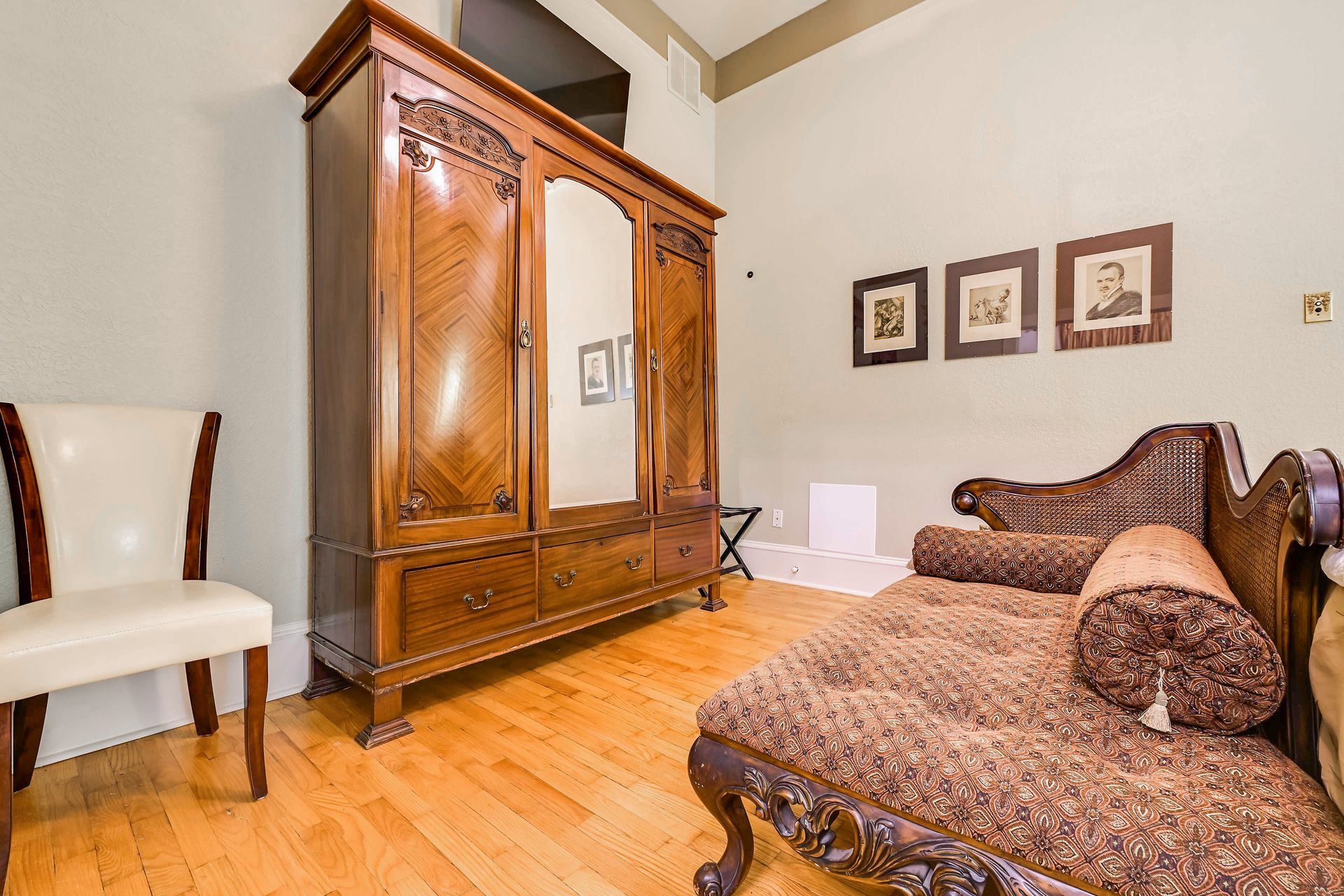 A bedroom with a wooden wardrobe, a bed with patterned bedding, a chair, and framed pictures on the wall.