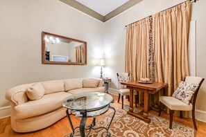 A living room with a beige sofa, a glass coffee table, a wooden dining table, a chair, a lamp, a mirror, and curtains. - Seclusion Suite- The Decatur Suites (Decatur)