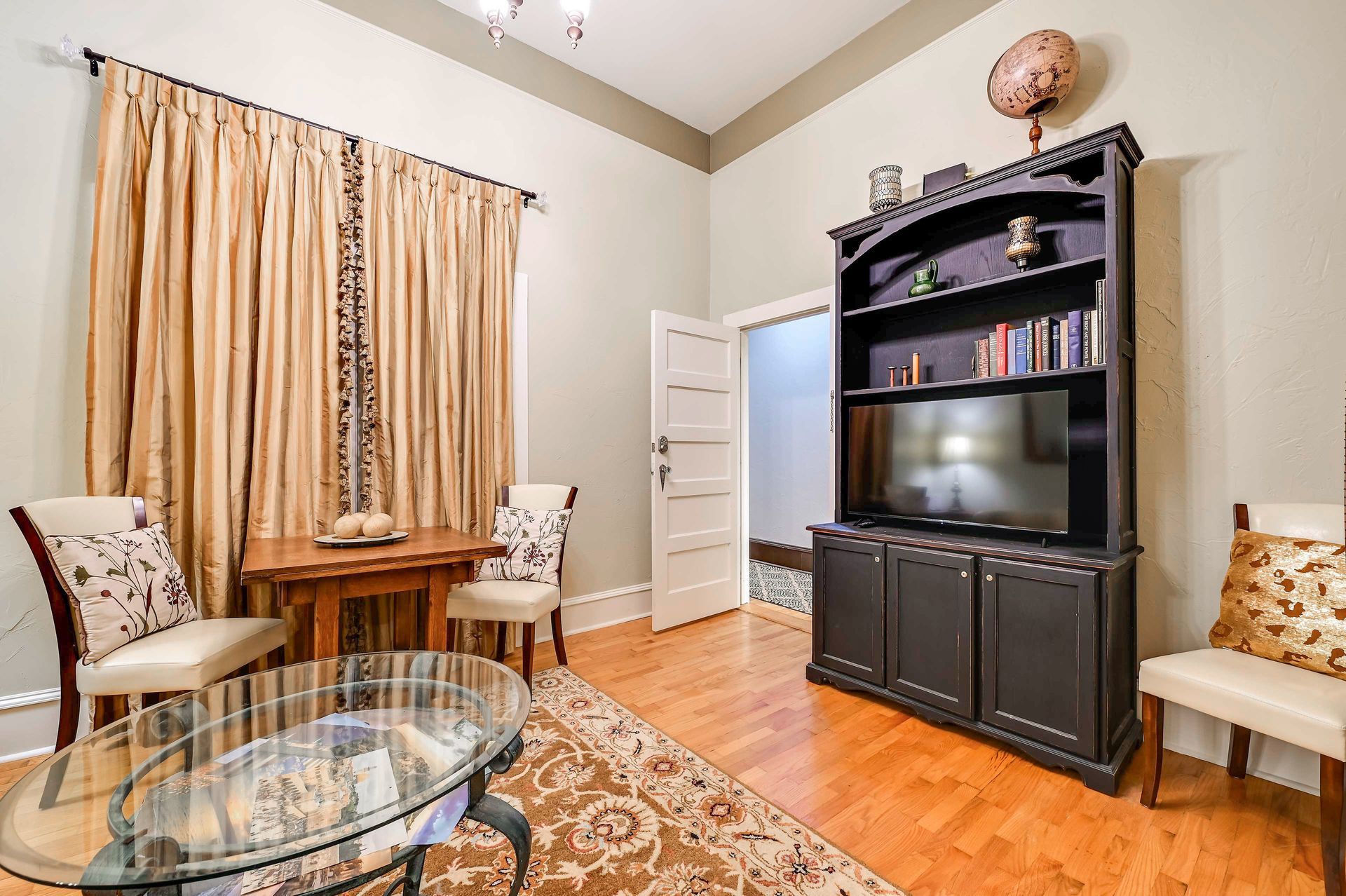 A living room with a glass coffee table, two chairs, a bookshelf, and a large window with curtains.