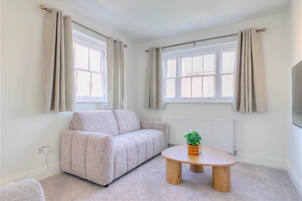 Ein Zimmer mit einer Couch, einem runden Holztisch und zwei Fenstern mit Vorhängen.