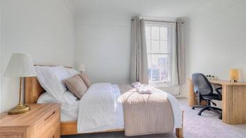 A bedroom with a bed, bedside table, lamp, desk, chair, and a window with curtains.