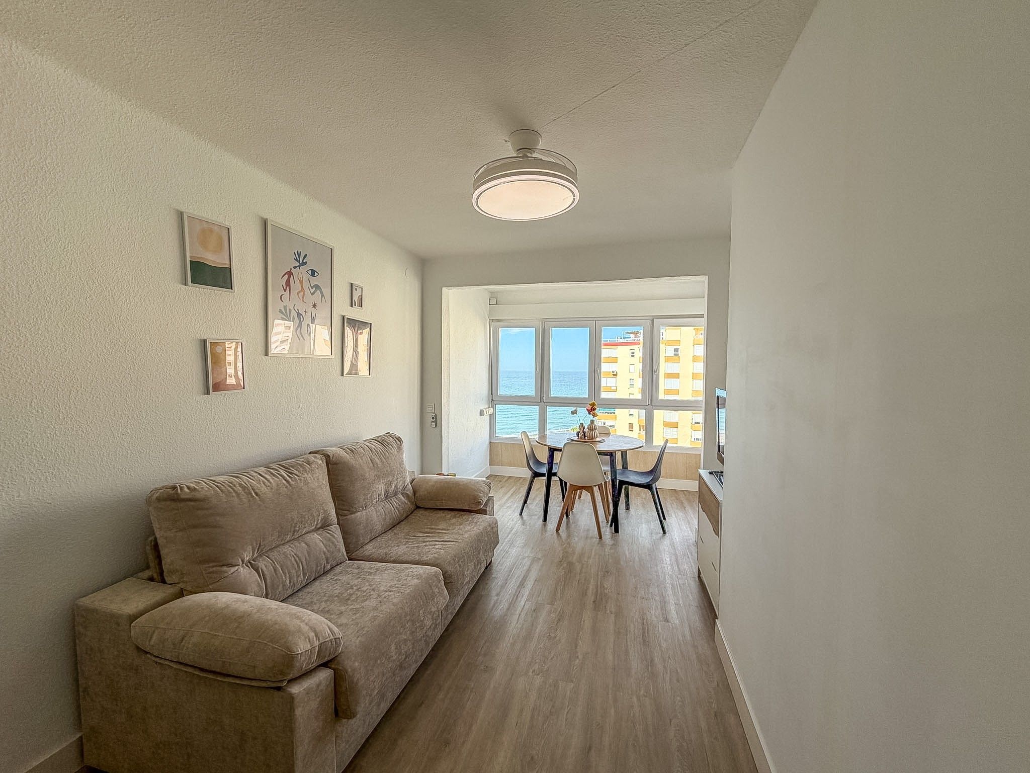 Een woonkamer met een beige bank, een eethoek met een tafel en stoelen, en een groot raam met uitzicht op gebouwen en de zee.