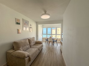 A living room with a beige sofa, a dining area with a table and chairs, and a large window with a view of buildings and the sea. - Apartment 'Apartamento en primera línea con vistas al mar en Torrox Costa' (Torrox)