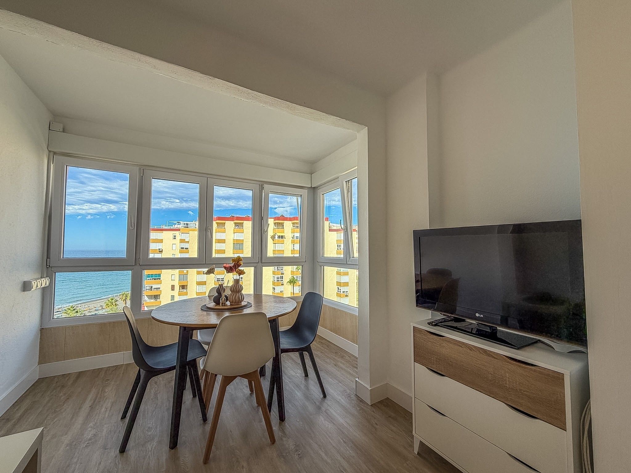Een kamer met een eettafel, stoelen, een televisie en grote ramen met uitzicht op gebouwen en een waterpartij.