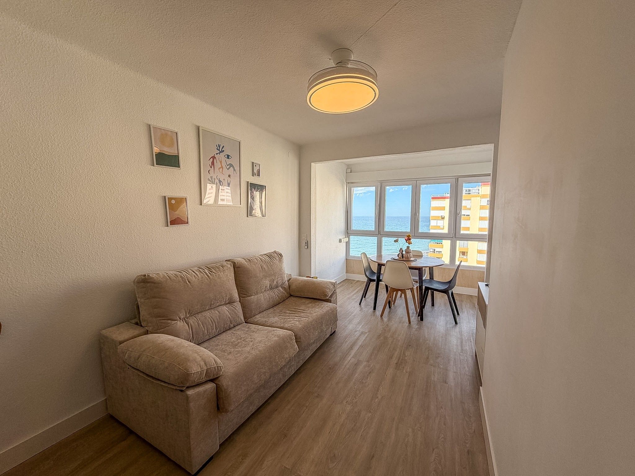Un salon avec un canapé beige, une table à manger avec des chaises, et une vue sur la mer grâce à de grandes fenêtres.